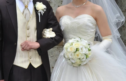 A bride in a white strapless gown holding a bouquet of white roses stands beside a groom in a dark suit and ivory vest.