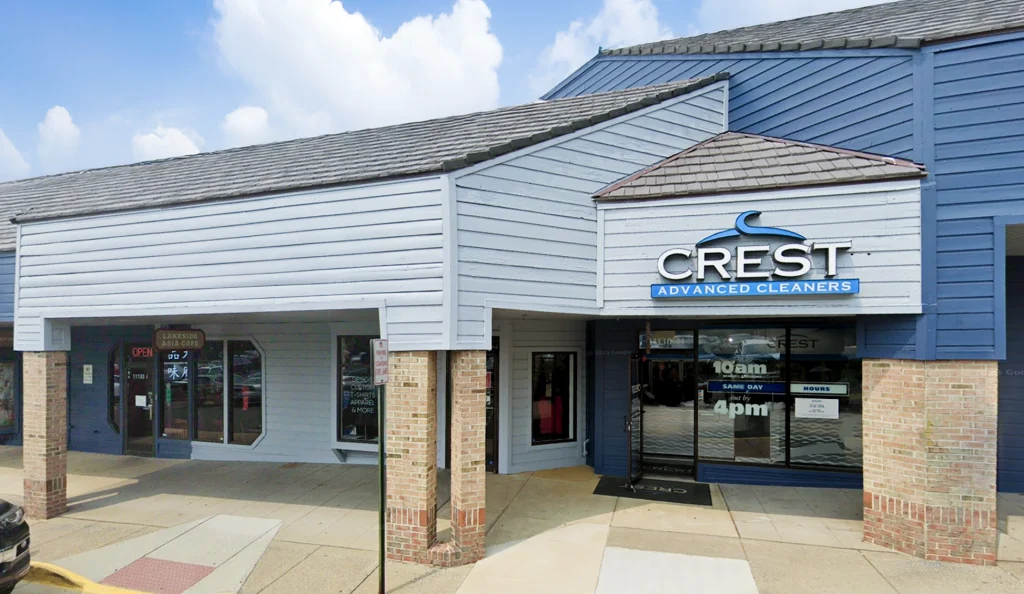A storefront for Crest Advanced Cleaners with a glass door displaying business hours, located in a shopping plaza with light blue siding and brick columns.