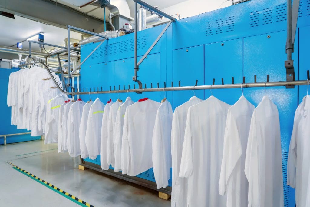 Rows of white lab coats hanging on a metal rack in front of large blue industrial laundry machines in a clean facility.