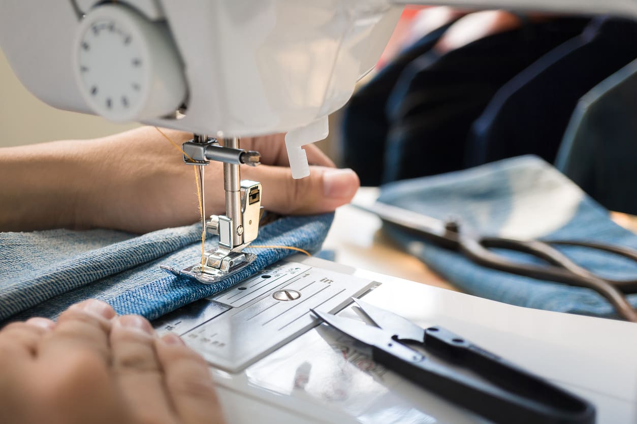 Close-up of hands using a sewing machine to stitch blue denim fabric, with sewing tools visible in the background.