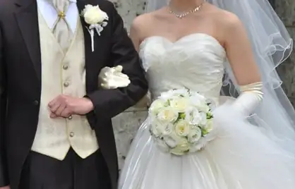 A bride in a white strapless wedding dress holding a bouquet of white roses stands beside a groom in a dark suit and ivory vest.